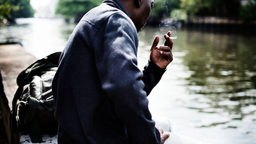 Person in smoking by the water with a backpack nearby.
